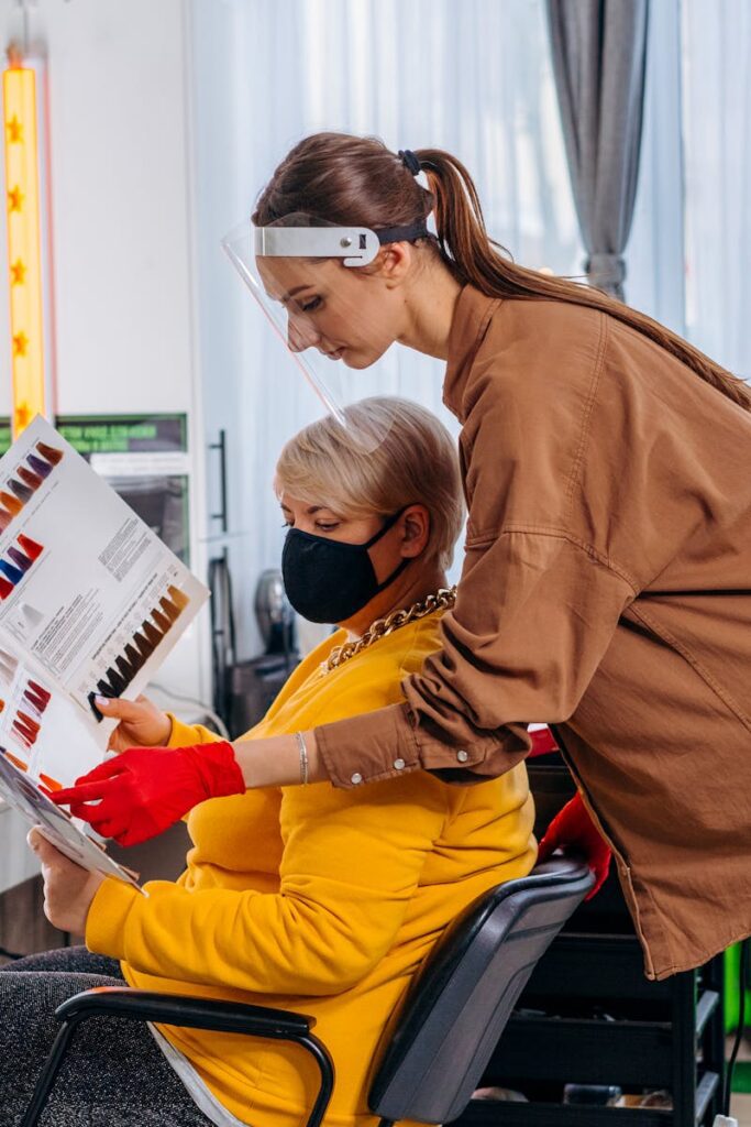 pexels-photo-7819723-7819723 Side view female master in medical mask and gloves showing album with hairstyles images to client on chair in salon