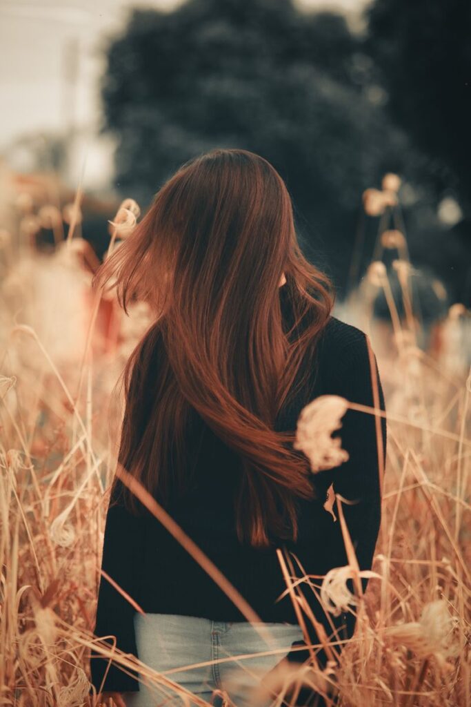 A woman with long brown hair stands in a grass field wearing a black outfit.