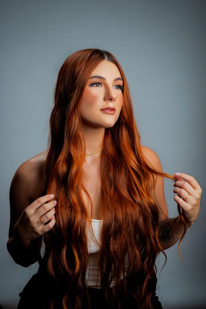 pexels-photo-15422149-15422149 Elegant portrait of a red-haired woman with long hair against a neutral gray background.