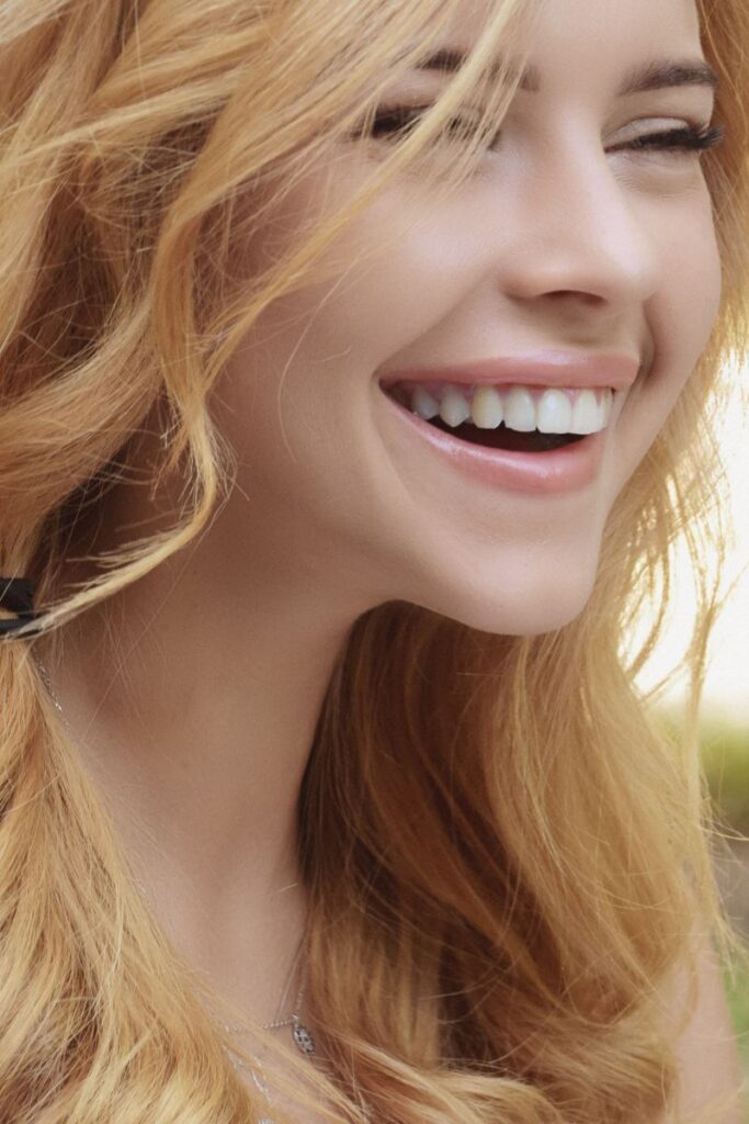 Close-up portrait of a joyful young woman with blonde hair smiling brightly outdoors.