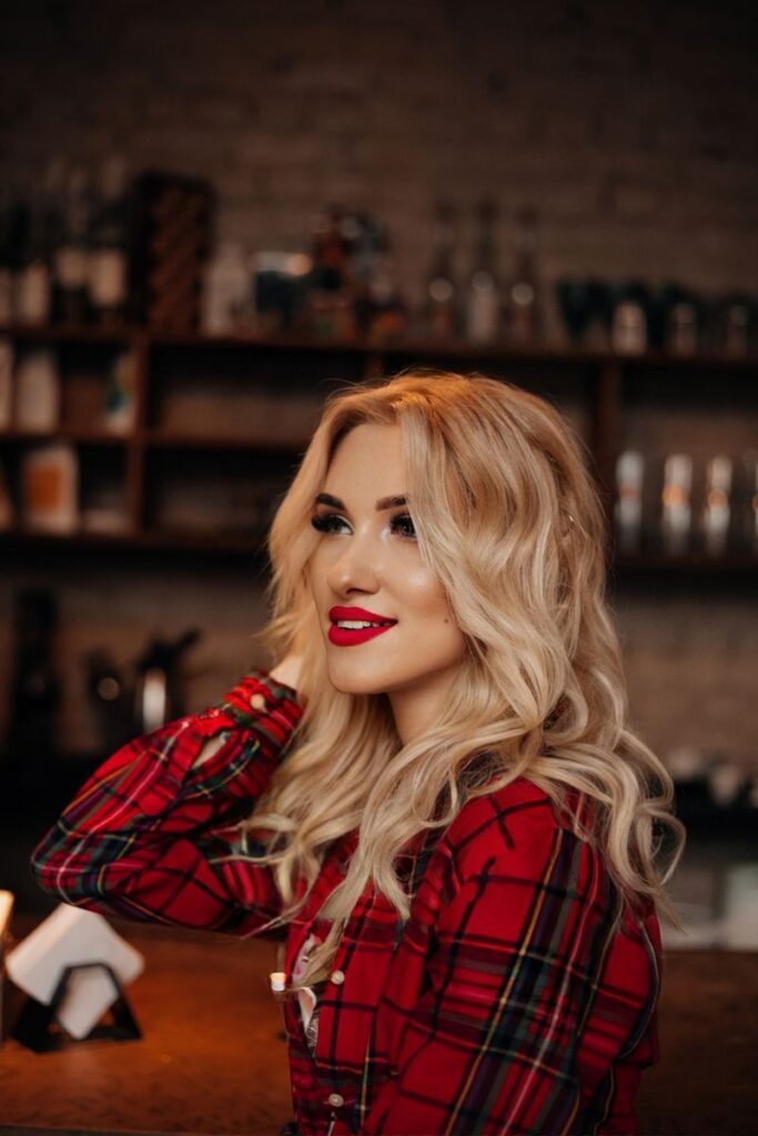 pexels-photo-20153934-20153934 Elegant blonde woman in a red plaid shirt smiling in a cozy indoor bar setting.