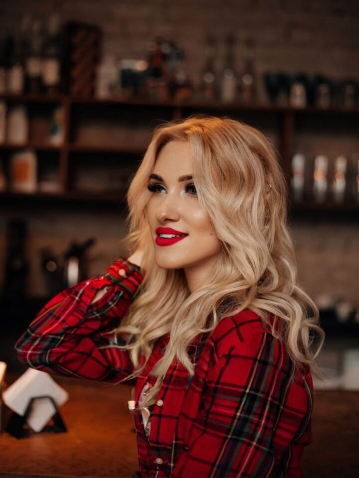 Elegant blonde woman in a red plaid shirt smiling in a cozy indoor bar setting.