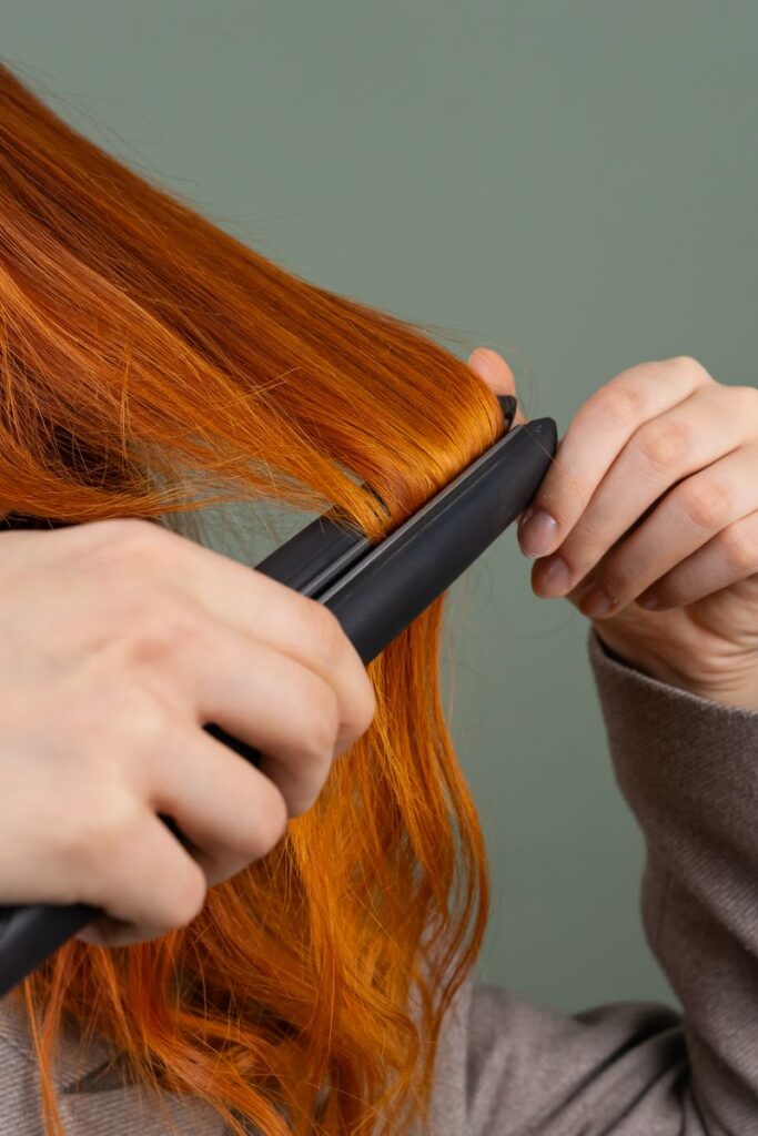 Close-up of a woman styling her red hair using a hair straightener against a green background.