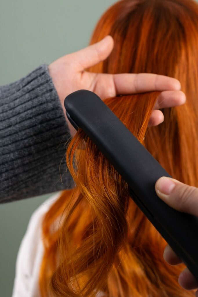 Hands styling long red hair using a flat iron indoors. Haircare and beauty concept.