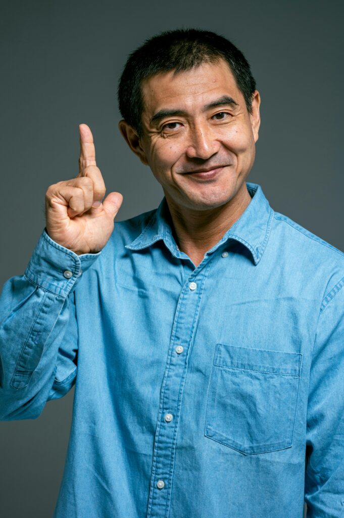 Portrait of an Asian man in a blue denim shirt, smiling and pointing upwards.