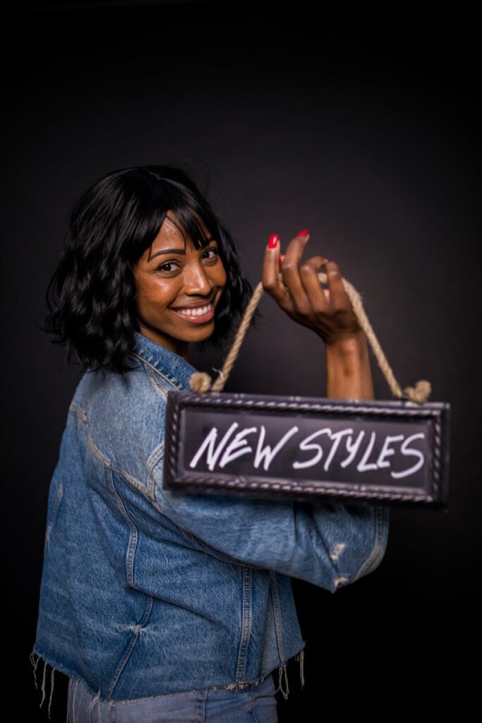 Portrait of a woman in denim jacket holding a 'New Styles' sign, smiling over her shoulder in a studio setting.