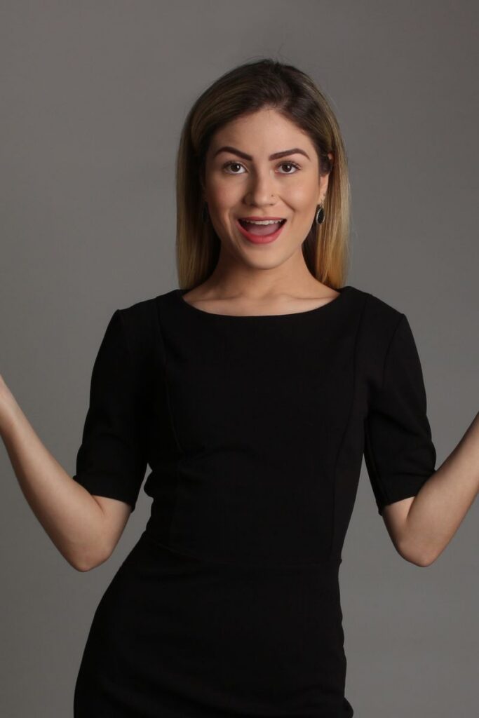 A woman in a black dress poses confidently with arms open in a studio setting.